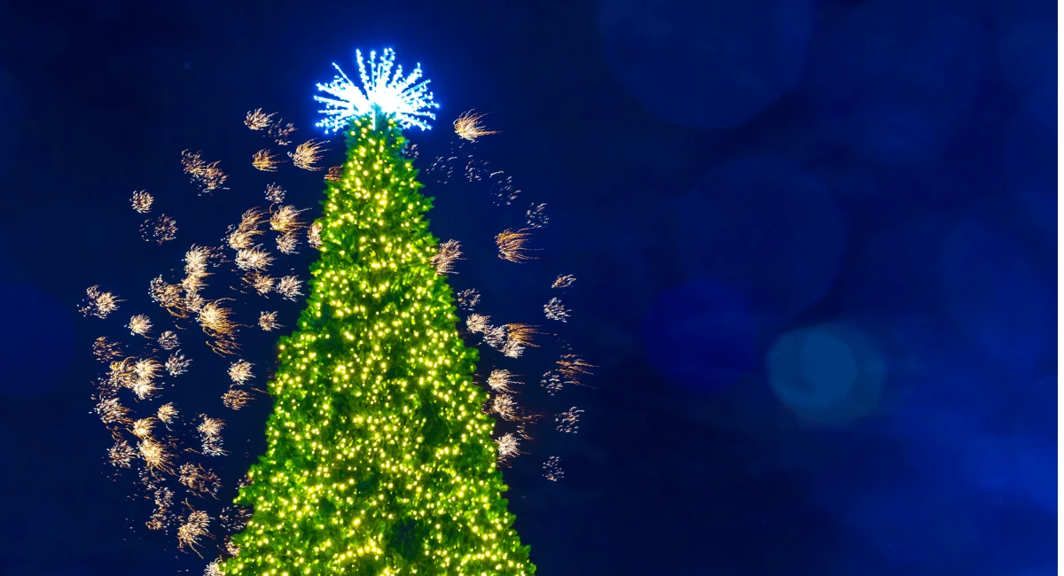 A brilliantly lit tree with a bright star on top and fireworks going off in the background under night skies.