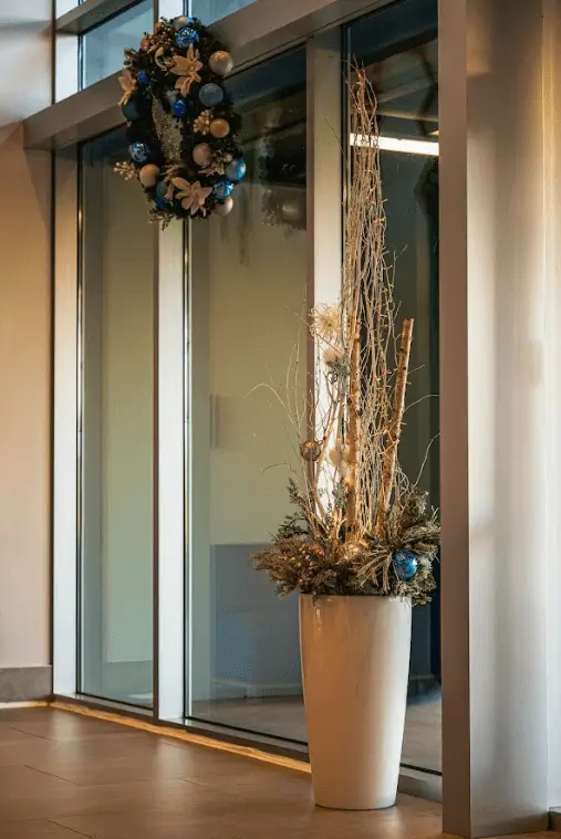A tall, slender floral arrangement in a white floor vase featuring birch branches, evergreen sprigs, and blue ornaments, positioned next to a glass wall.