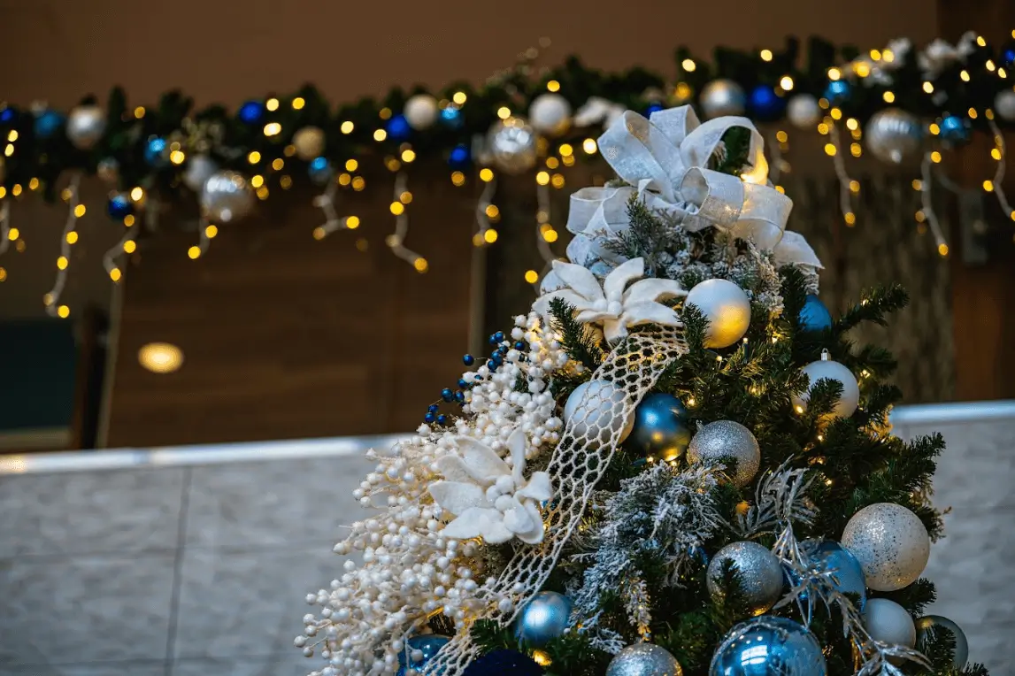 Close-up of the top of a Christmas tree decorated with a large white ribbon, white poinsettias, and blue and silver ornaments against a blurred background of festive lights.