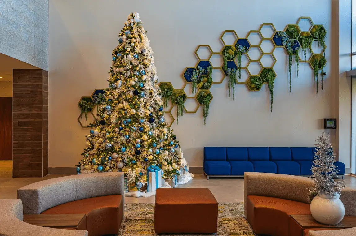 A large, brightly lit Christmas tree decorated with blue, silver, and white ornaments, standing in a lobby next to a geometric honeycomb wall installation.