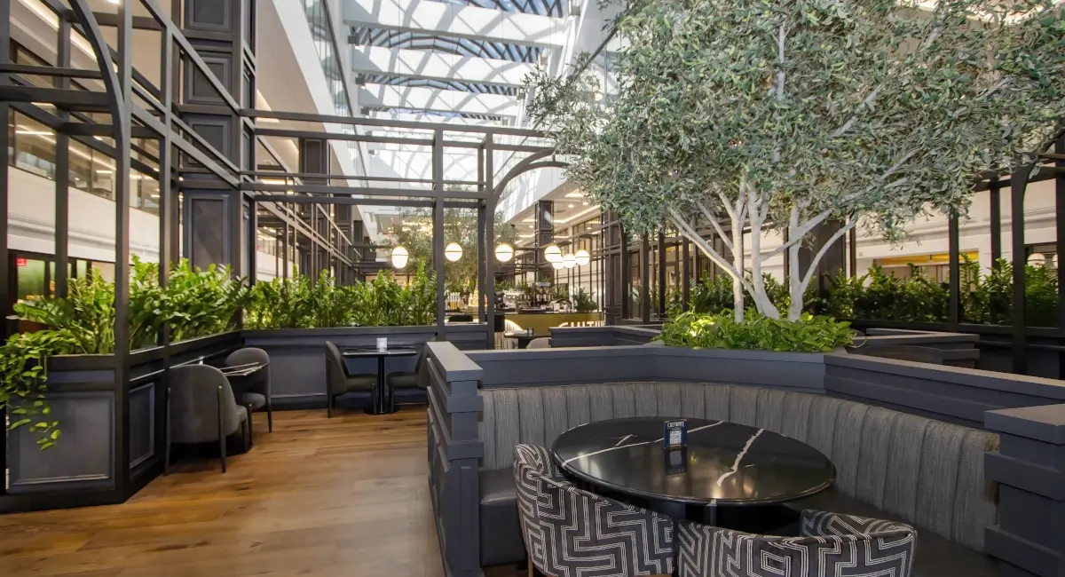 Upscale office lounge seating area featuring indoor trees, booth seating, and a glass-ceiling atrium.