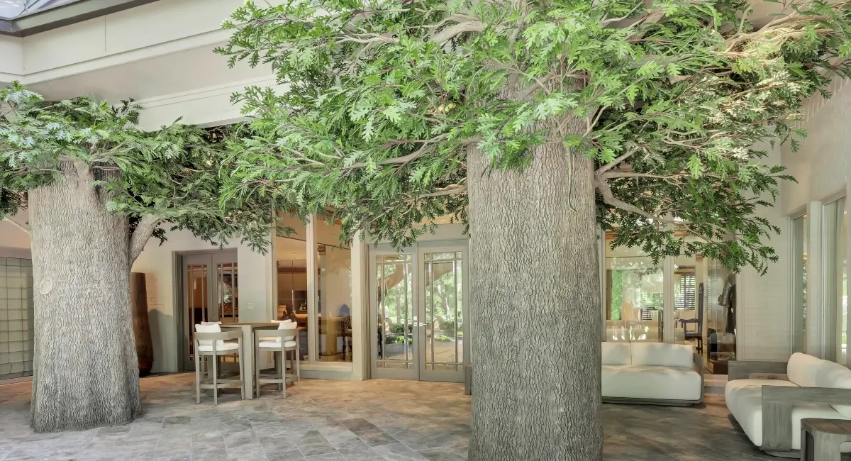 Close-up of large-scale artificial trees providing a leafy canopy over a modern stone patio lounge area.
