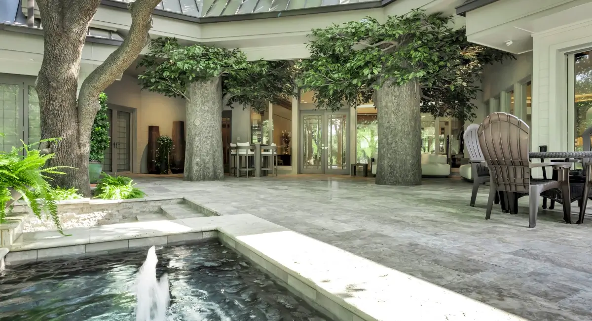A spacious outdoor courtyard with a stone water fountain, patio seating, and large artificial oak trees.