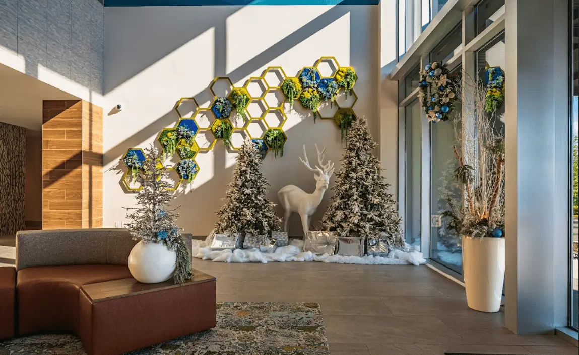 A modern lobby decorated for the holidays with a large white reindeer statue, flocked Christmas trees, and a honeycomb-patterned wall feature with greenery and blue accents.