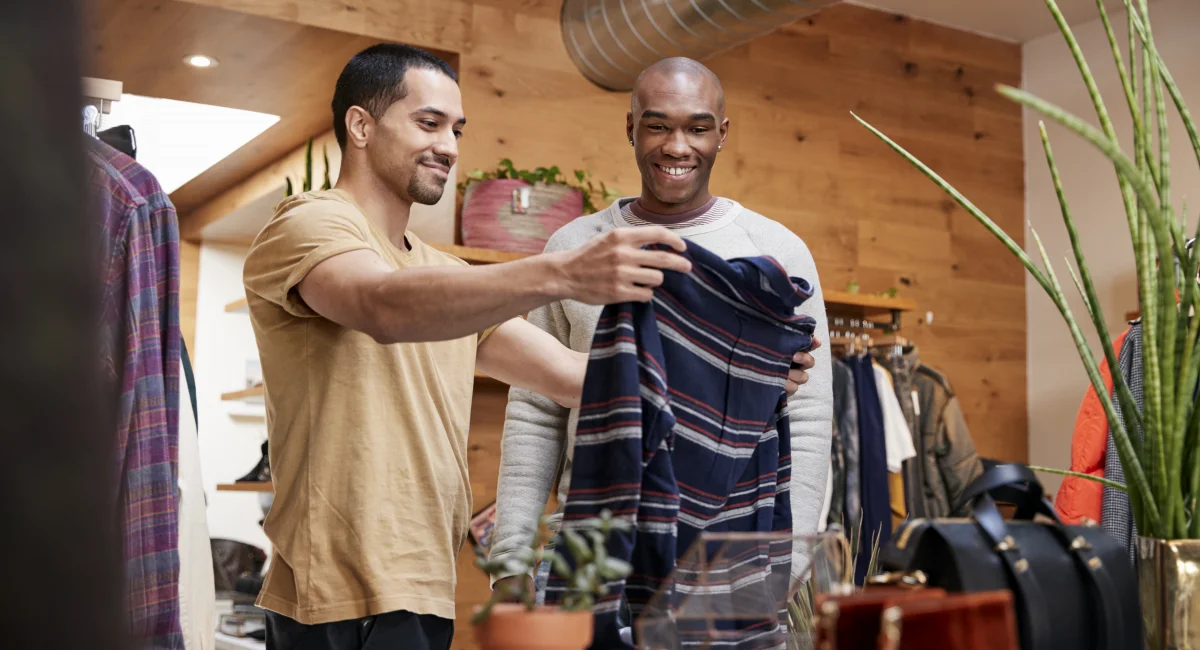 image-banner-stock-two-young-men-holding-up-clothes-to-look-at-in-901866822.webp